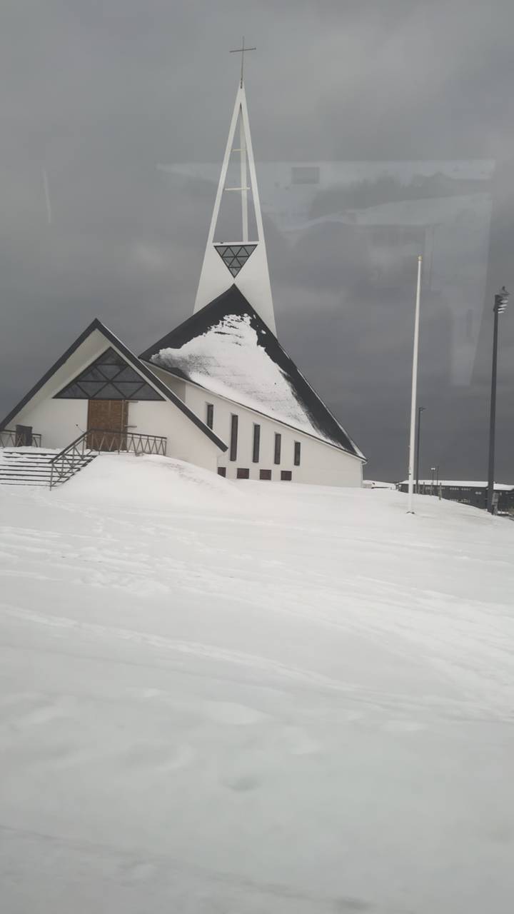 Modern white church with steep black roof blanketed in snow against a brooding sky.