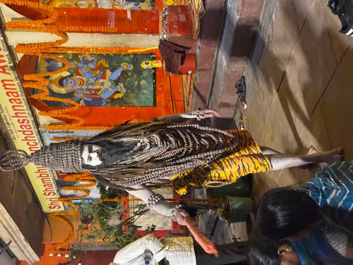 Ash-smeared holy man dressed as Lord Shiva with dreadlocks and rudraksha beads inside a decorated shrine.