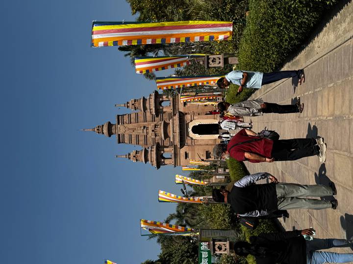 Visitors walk a flag-lined path toward a sandstone Buddhist temple under bright blue sky.
