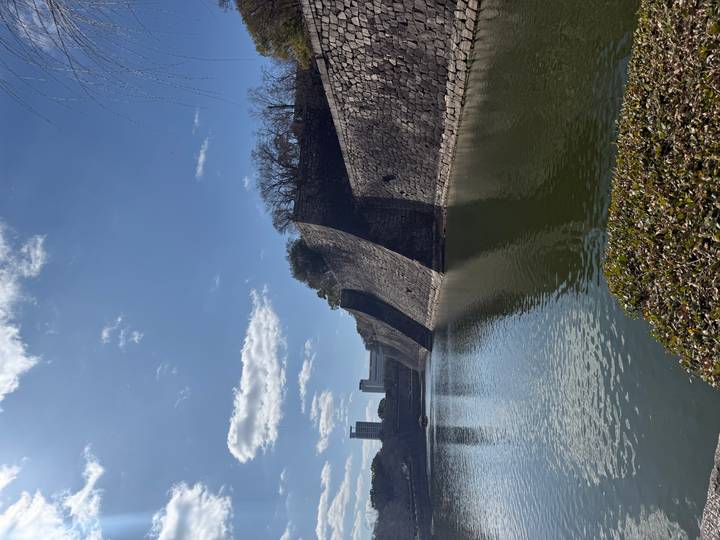 Quiet moat surrounded by high stone ramparts with city skyline peeking in the distance under clear skies.