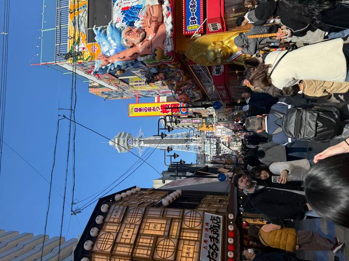 Crowded pedestrian street bursting with colorful shopfronts leading to a towering Tsutenkaku observation tower.