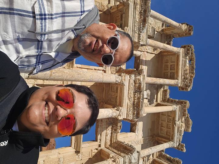 Smiling travellers take a close-up selfie with the ancient limestone columns of the Library of Celsus behind them.