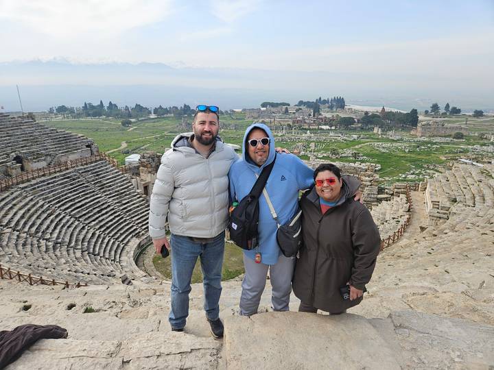 Three friends stand on the stone tiers of Hierapolis amphitheatre overlooking green plains and distant mountains.