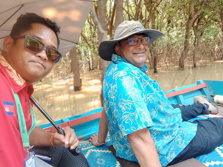 Two men in a small blue wooden boat navigating a flooded forest canal, one taking a selfie.