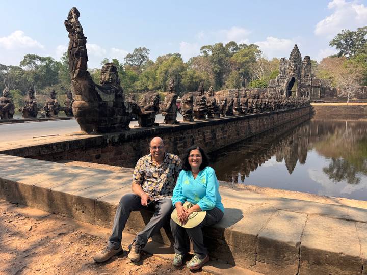 Couple seated beside the moat of Angkor Thom with a row of guardian statues and gate in view.