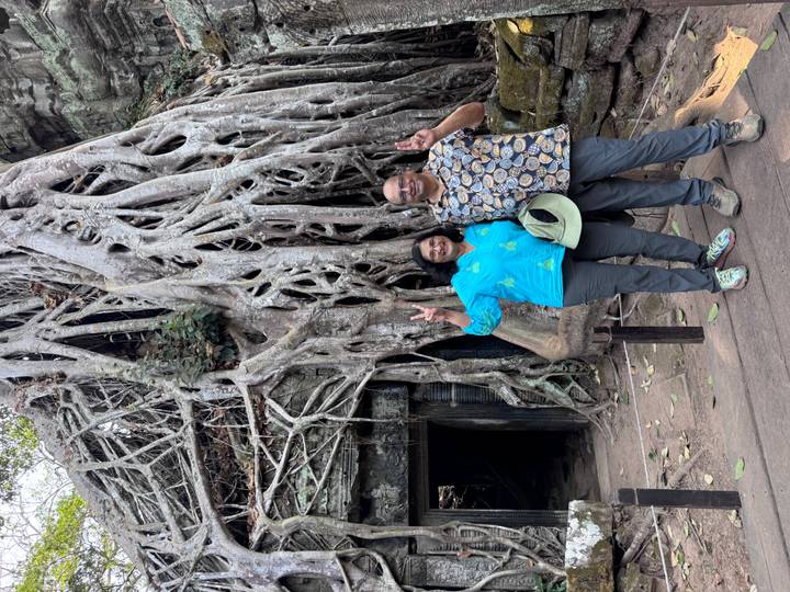 Couple flashing peace signs beneath massive tree roots engulfing ruins at Ta Prohm temple.