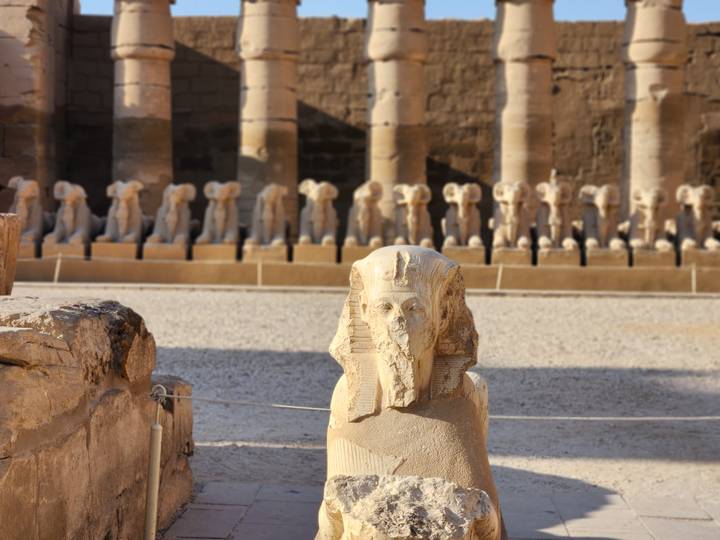 A ram-headed sphinx statue stands in sharp focus with colonnaded rows of statues blurred behind at Karnak Temple.