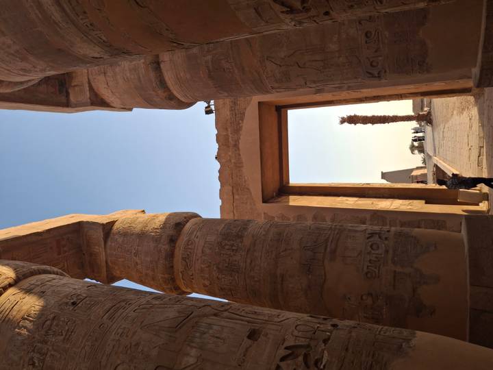 Tall engraved sandstone columns framing a doorway that looks out toward an obelisk and palm in an ancient Egyptian temple.