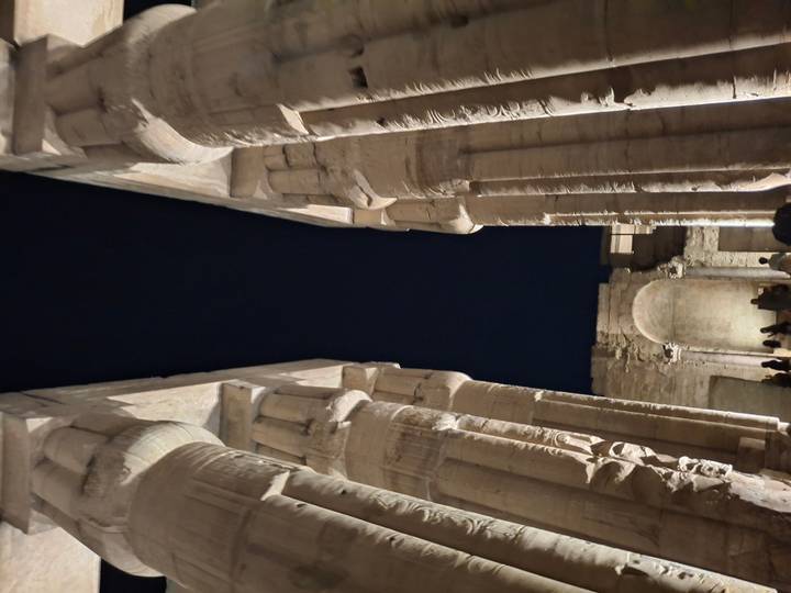 Night-time view between massive lit columns inside an Egyptian temple, with visitors silhouetted at the base.