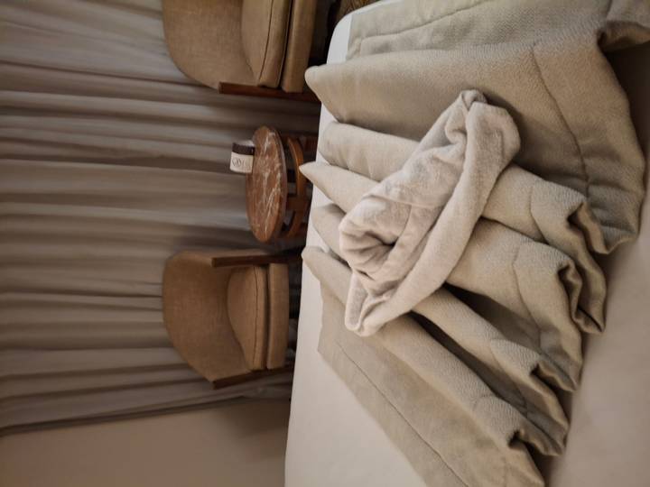 Hotel or cruise-ship cabin with folded beige towels on the bed, two chairs and a small round table in soft lighting.