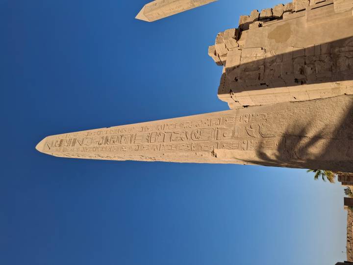Tall stone obelisk covered in hieroglyphs standing under a bright blue sky in an Egyptian temple complex.