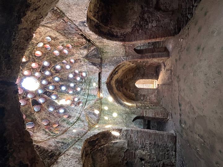 Ancient domed bathhouse interior with circular skylights projecting beams of sunlight onto rough stone walls.