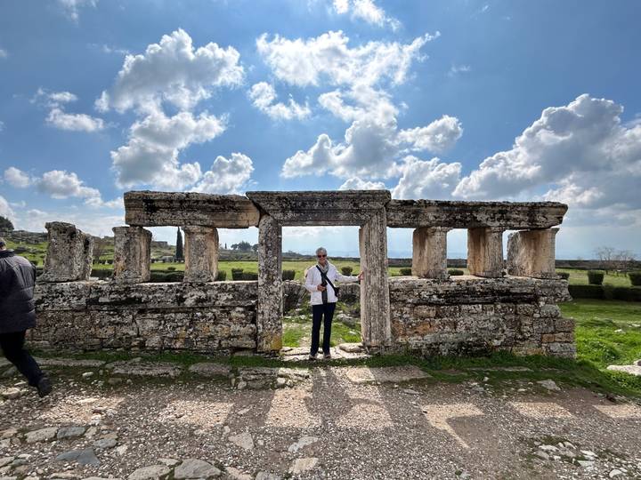 Traveler stands framed by ancient stone columns and architrave ruins beneath a sky dotted with clouds.
