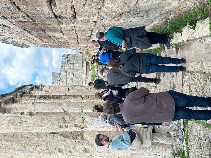 Tour group listens to a guide while standing between towering ancient limestone columns and ruins.