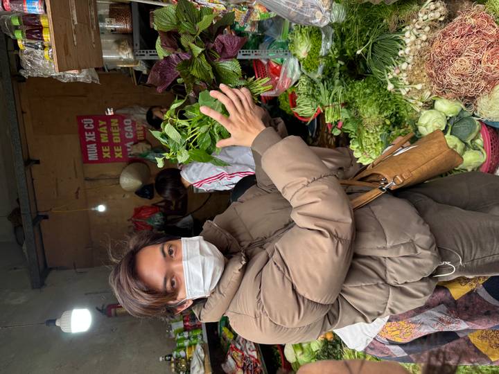 Masked guide holding bunches of fresh herbs while explaining their uses at a market stall.