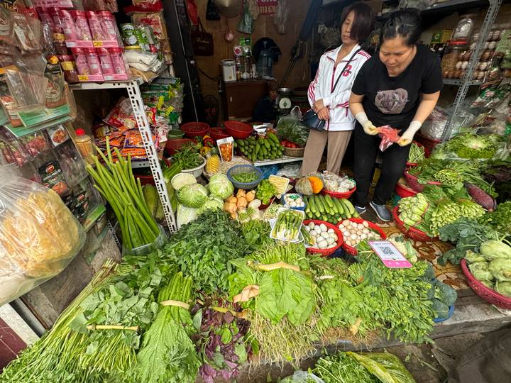 Heaped piles of vegetables, gourds and herbs fill a cramped street stall while shoppers browse.