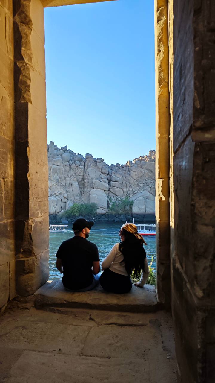 Two travellers framed by ancient stone doorway admire rocky Nile islands and passing boat under clear blue sky.