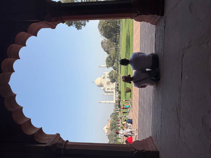 Couple sitting under an arched pavilion gazing at the distant Taj Mahal across manicured lawns.