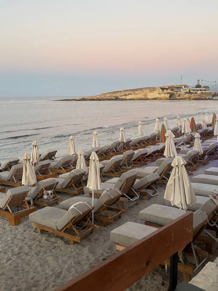 Neat rows of empty sun loungers and umbrellas line a calm sandy beach at sunset.