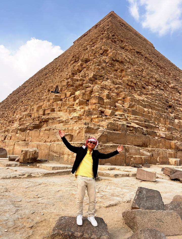 Visitor poses with arms raised at the massive stone base of a pyramid.