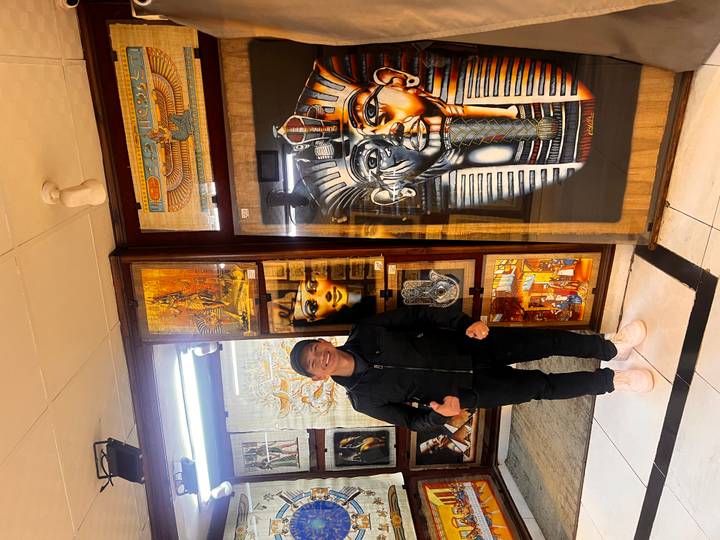 Visitor gives thumbs-up beside colourful papyrus artwork in an Egyptian store.