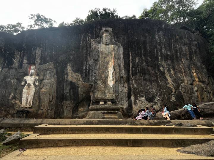 Ancient rock cliff with large standing Buddha statues carved into the stone, several visitors sitting at the base.