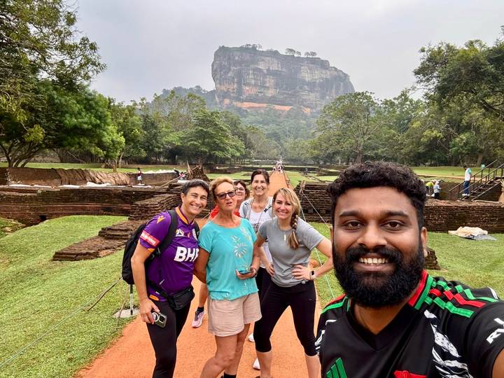 Selfie group smiles with lush ruins and Sigiriya rock fortress behind them.