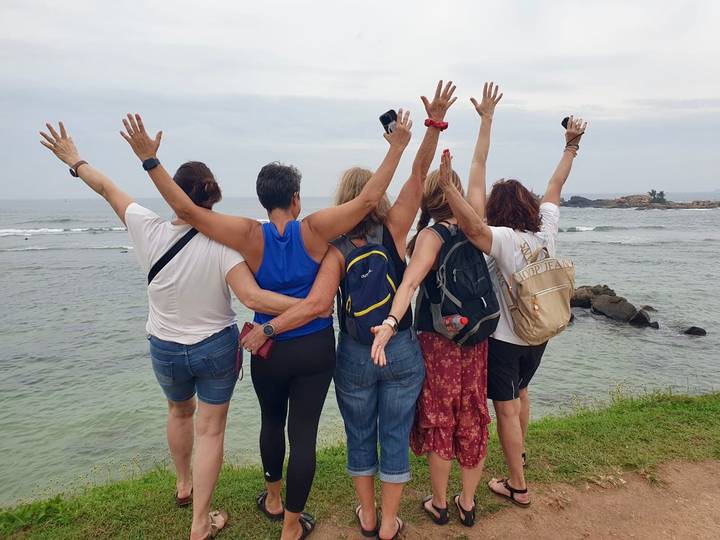 Five women stand with arms raised facing the sea on a rocky shoreline.