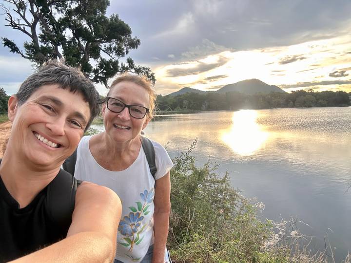 Two women smile for a selfie beside a reflective lake at sunset with mountains behind.