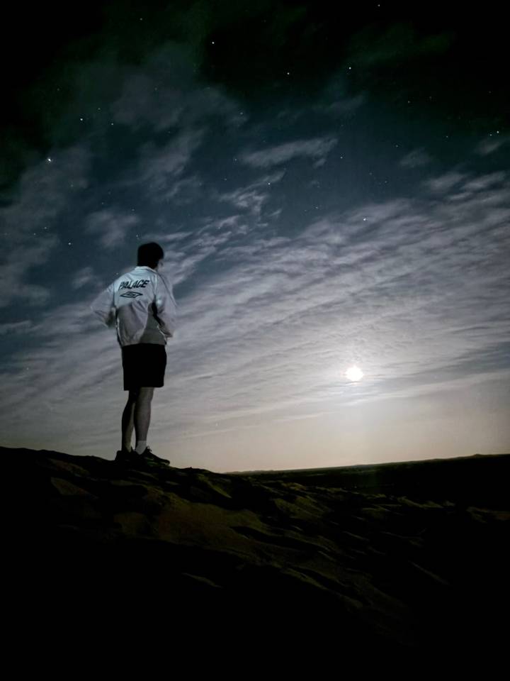 Person standing on a ridge at night looking out at a bright moon and streaked clouds.