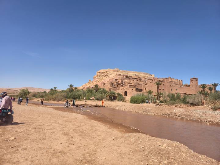 View of the earthen fortified village of Ait Benhaddou across a shallow river with tourists nearby.