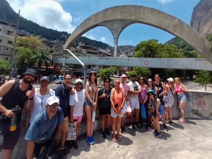 Large tour group posing beneath a concrete arch with a hillside favela in the background.