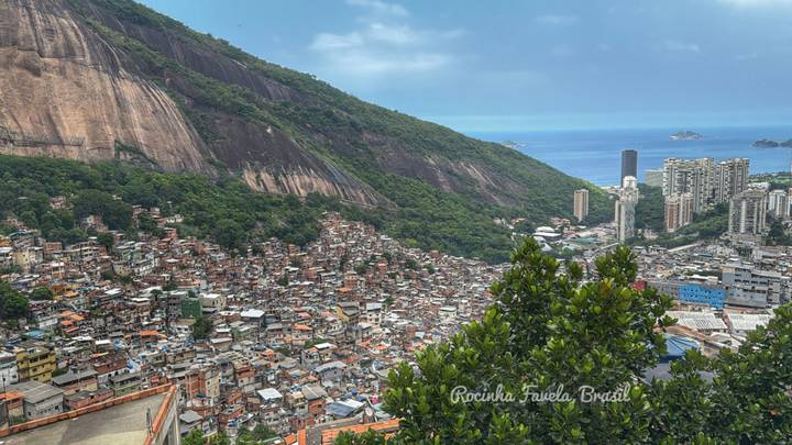 Panoramic view of Rocinha favela with ocean and city towers beyond, text label overlaid at bottom.