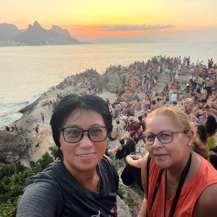 Two women selfie with a huge crowd gathered on a rocky point overlooking the sea at sunset.