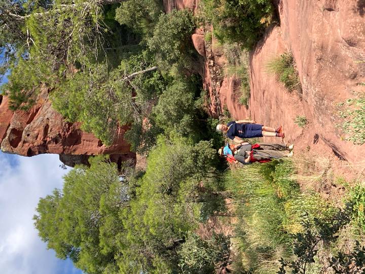 Small hiking party walks along red sandstone trail beneath towering cliff and pine trees in Catalonia