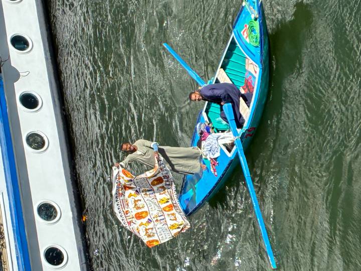 Two vendors in small boat hold up colourful textiles to cruise passengers on the Nile