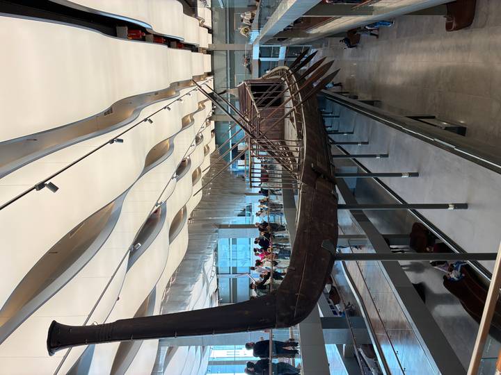 Ancient wooden solar boat displayed in large modern gallery with crowds viewing from balconies