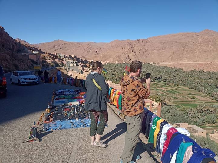Tourists photographing colourful roadside handicraft stalls overlooking lush oasis valley in Morocco