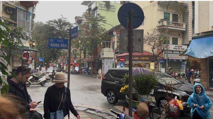 Bustling Hanoi street with scooters, shops and pedestrians during a light drizzle.