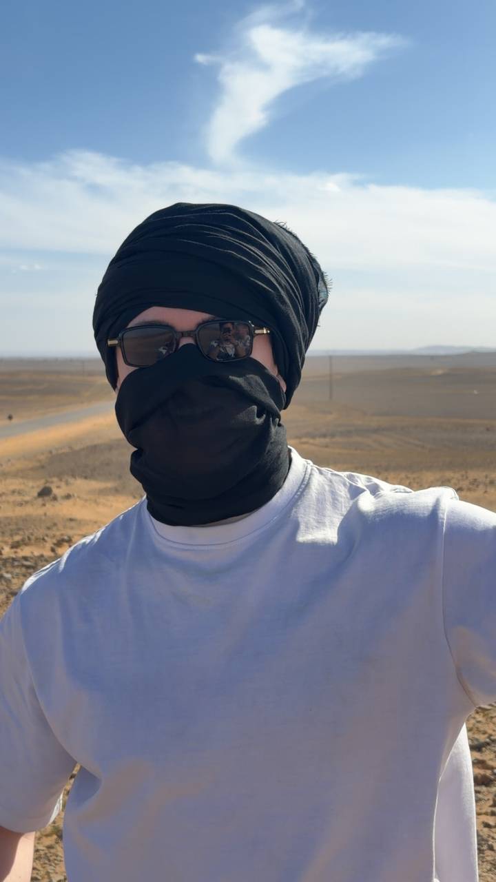Close-up portrait of a traveler wearing sunglasses and a desert head scarf with sandy plains behind.