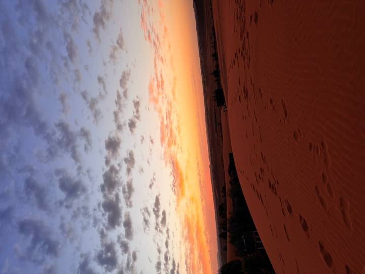 Footprints lead across red dunes toward a vibrant sunrise with pastel clouds.