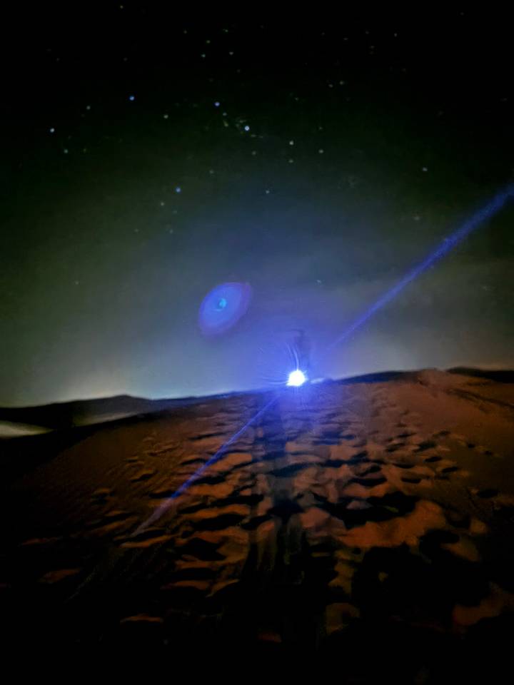 A blurry night shot of desert sand with a single bright flashlight beam creating lens flare.