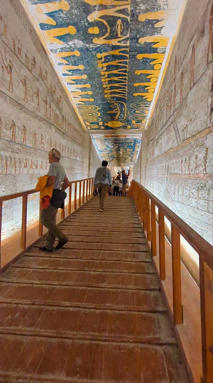 Tourists walk along a wooden ramp inside a hieroglyph-covered tomb corridor illuminated by artificial light.