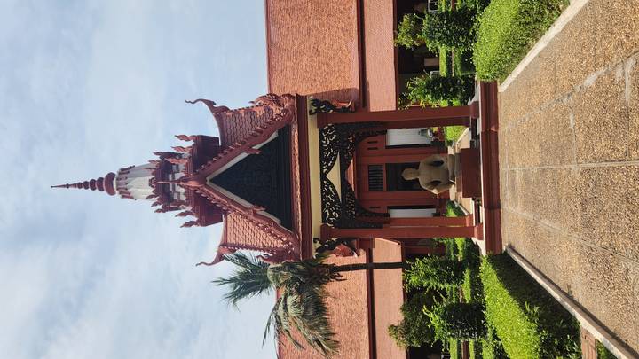 Ornate Khmer-style pavilion with red tiled roof and a seated Buddha statue in a manicured courtyard under a bright blue sky.