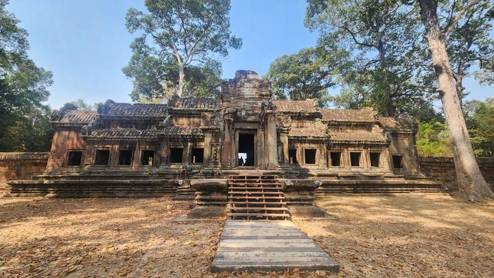 Stone temple ruin nestled among tall trees with a central staircase leading to multiple windowed chambers, carpeted by dry leaves.