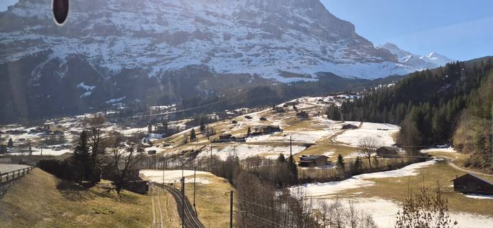 Snow-patched alpine valley with scattered chalets and railway tracks beneath the towering Eiger massif.