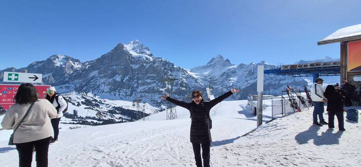Happy traveler spreads arms on a snowy summit with dramatic alpine peaks and ski lifts in the background.