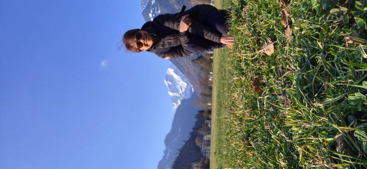 Traveler kneels in a grassy meadow with snow-capped peaks and forested slopes rising in the distance.