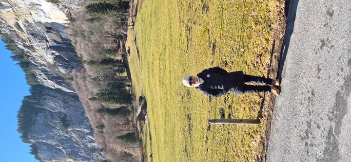Visitor in winter coat stands beside a pasture backed by steep rocky cliffs and mixed conifer forest.