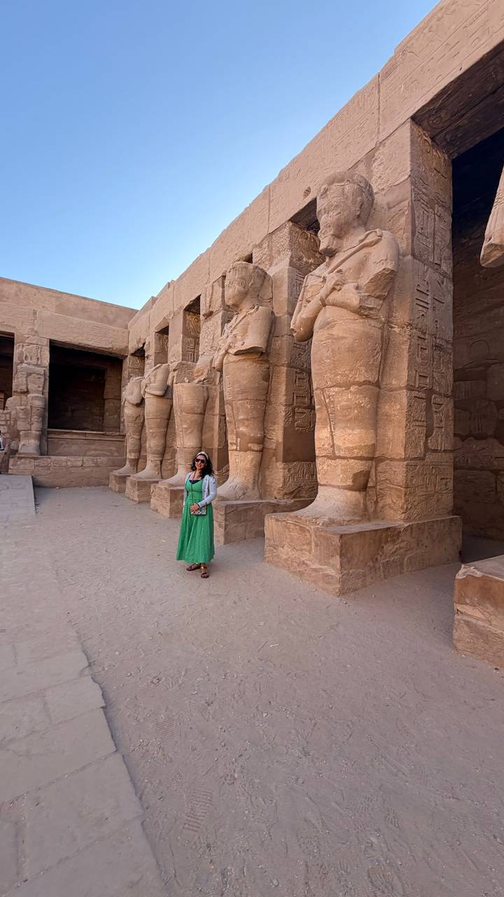 Woman standing among colossal sandstone statue legs in an ancient Egyptian temple.